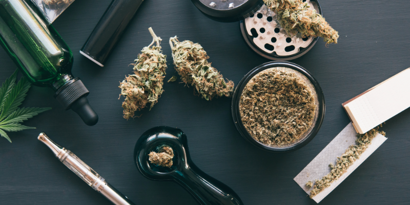 A variety of cannabis products and gear, including a tincture bottle, vape pen, flower, pipe, unrolled joint, and herb grinder, laid out on a table.