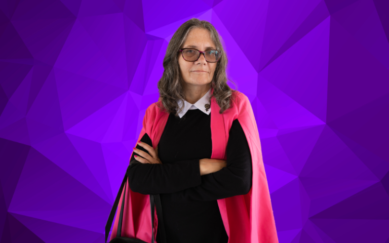 Coleen Cusack stands with her arms cross in a bright pink cape standing in front of a geometric purple background.