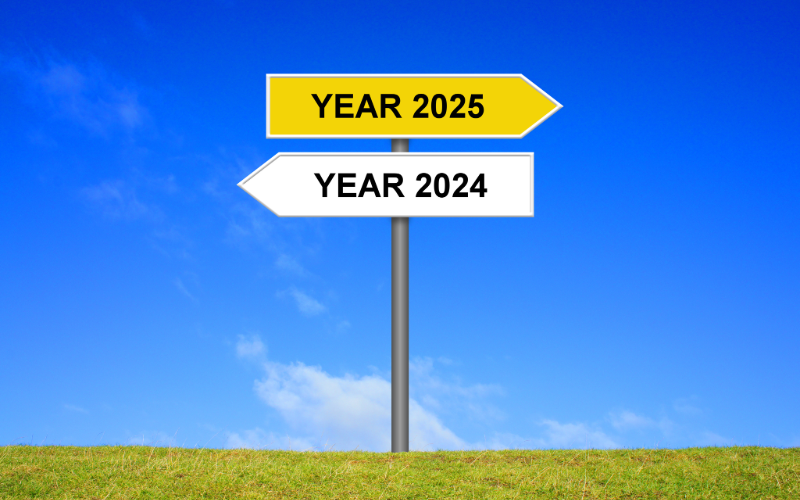 A sign is pictured on a lawn, with the upper portion pointing right with text reading YEAR 2025 and the bottom portion pointing left with text reading YEAR 2024